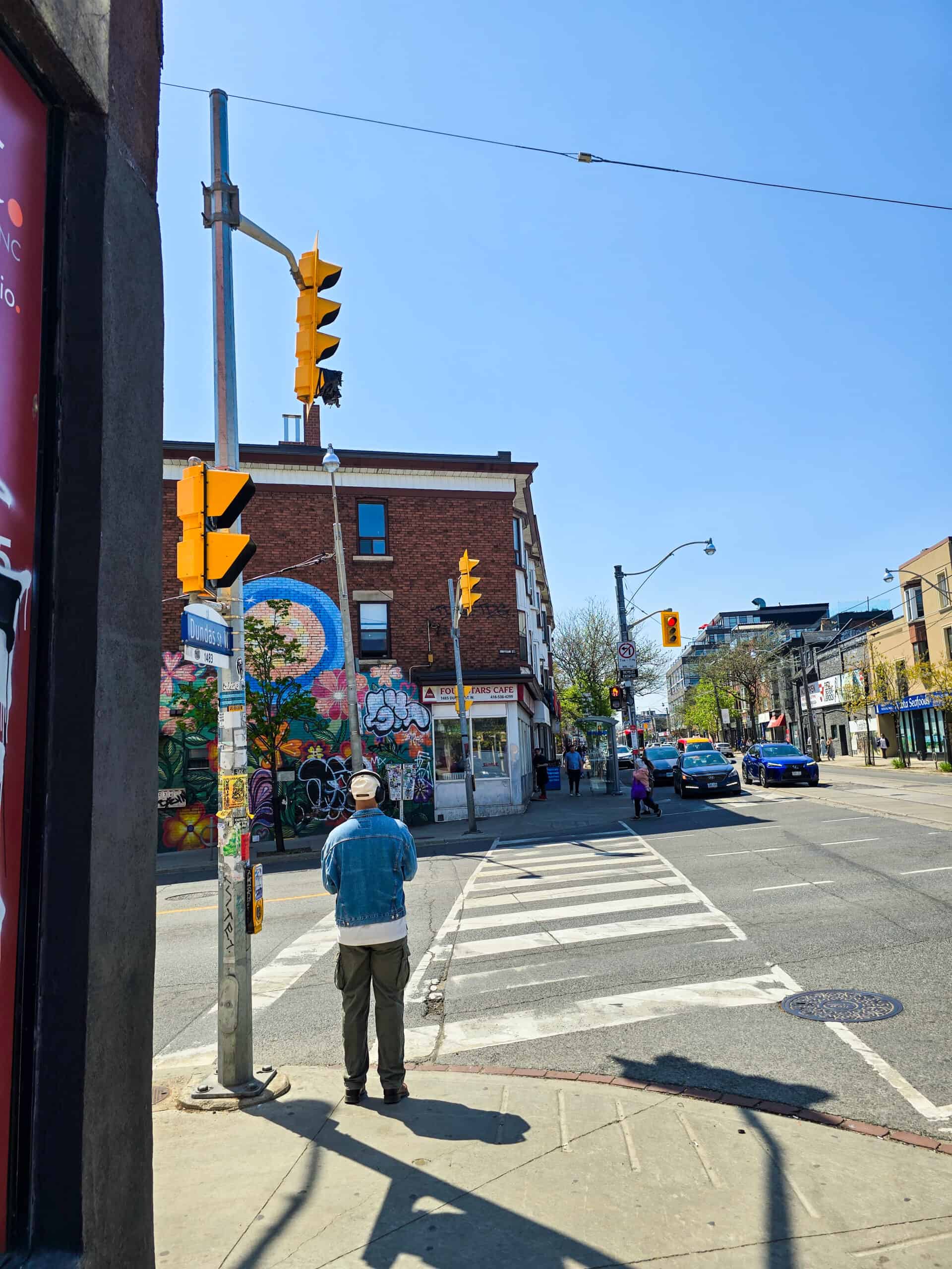 A man wearing headphones stands on a street corner in Toronto's Little Portugal with street art in front of him.